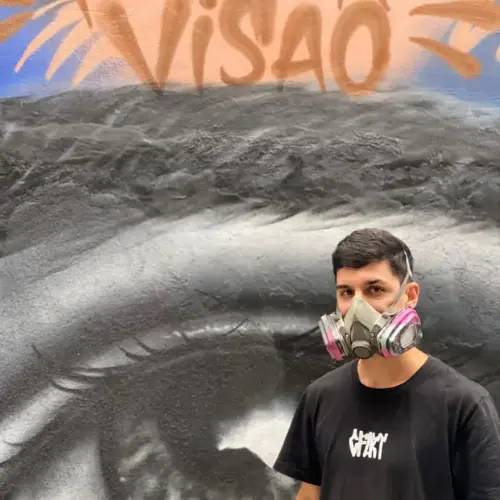 Artista com máscara de pintura e camisa preta. Ao fundo, a pintura de um olho e a palavra 'visão'.
