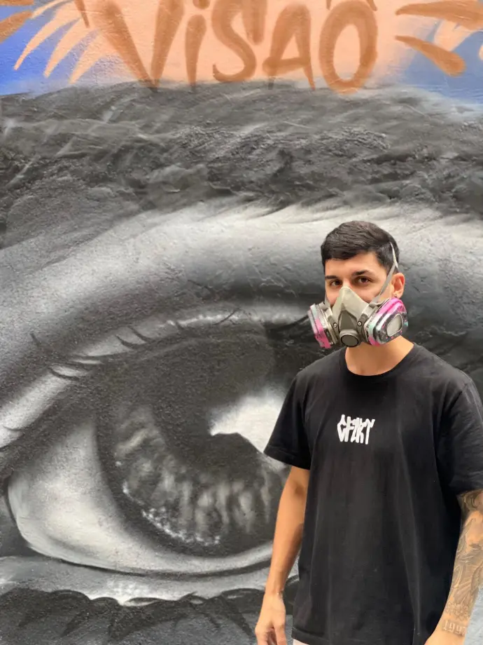 Artista com máscara de pintura e camisa preta. Ao fundo, a pintura de um olho e a palavra 'visão'.