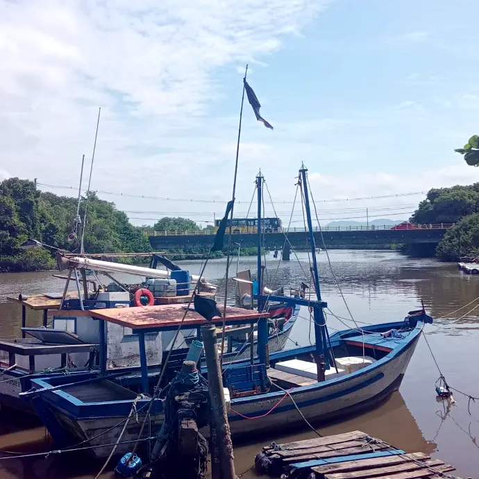 Barcos de pesca atracados à beira do rio com ponte ao fundo em fim de tarde ensolarado.
